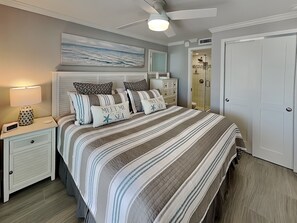 A bedroom with a bed, nightstands, a dresser, a ceiling fan, a painting, and a bathroom door. - Pointe South 101 | Stunning Views + Pool (Fort Myers Beach)
