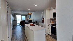 A modern kitchen with white cabinetry, a central island, and a dining area with a sofa and table. - Pointe South 306 | Modern Gulf Escape + Pool (Fort Myers Beach)