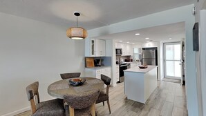 A modern kitchen with white cabinets, a central island, and a dining area with a round table and chairs. - Pointe South 306 | Modern Gulf Escape + Pool (Fort Myers Beach)