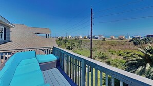 A wooden deck with blue cushions, a view of a beach, and houses in the background. - Prince Paradise | Spacious, Gulf Views, + Community Pool (Galveston)