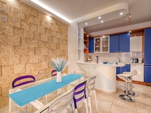 A modern kitchen with a stone backsplash, blue cabinets, and a glass dining table with purple chairs.