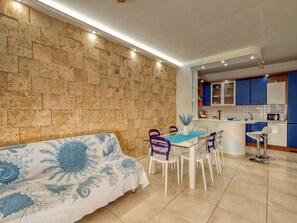 A modern kitchen with blue cabinets, a dining area with a glass table and purple chairs, and a stone-patterned wall.