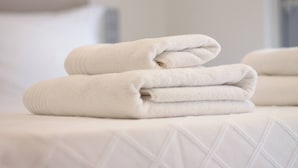 Uma toalha branca e bem dobrada sobre uma cama com colcha branca texturizada.