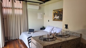 A hotel room with a bed, a dining table set with glasses and plates, an air conditioner, a painting on the wall, and a curtain.