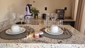 A dining table set with plates, cups, cutlery, and wine glasses, with a kitchen in the background.