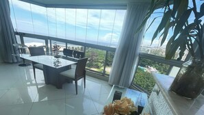 A modern dining area with a glass table and chairs, a vase of flowers, and a balcony with a view of the ocean and greenery. - Charming 2-bedroom Apartment With Ocean View in Rio (Rio de Janeiro)