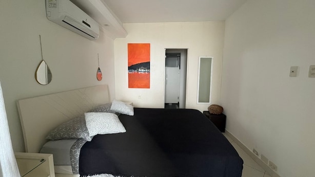 A bedroom with a bed, a bedside table, a wall-mounted air conditioner, a door, and a window. - Charming 2-bedroom Apartment With Ocean View in Rio (Rio de Janeiro)