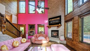 A cabin interior with a pink wall, a fireplace, a ceiling fan, and a sign for 'Broken Bow Cabin Rentals.' - Pretty in Pink: Sleeps 14 (Broken Bow)