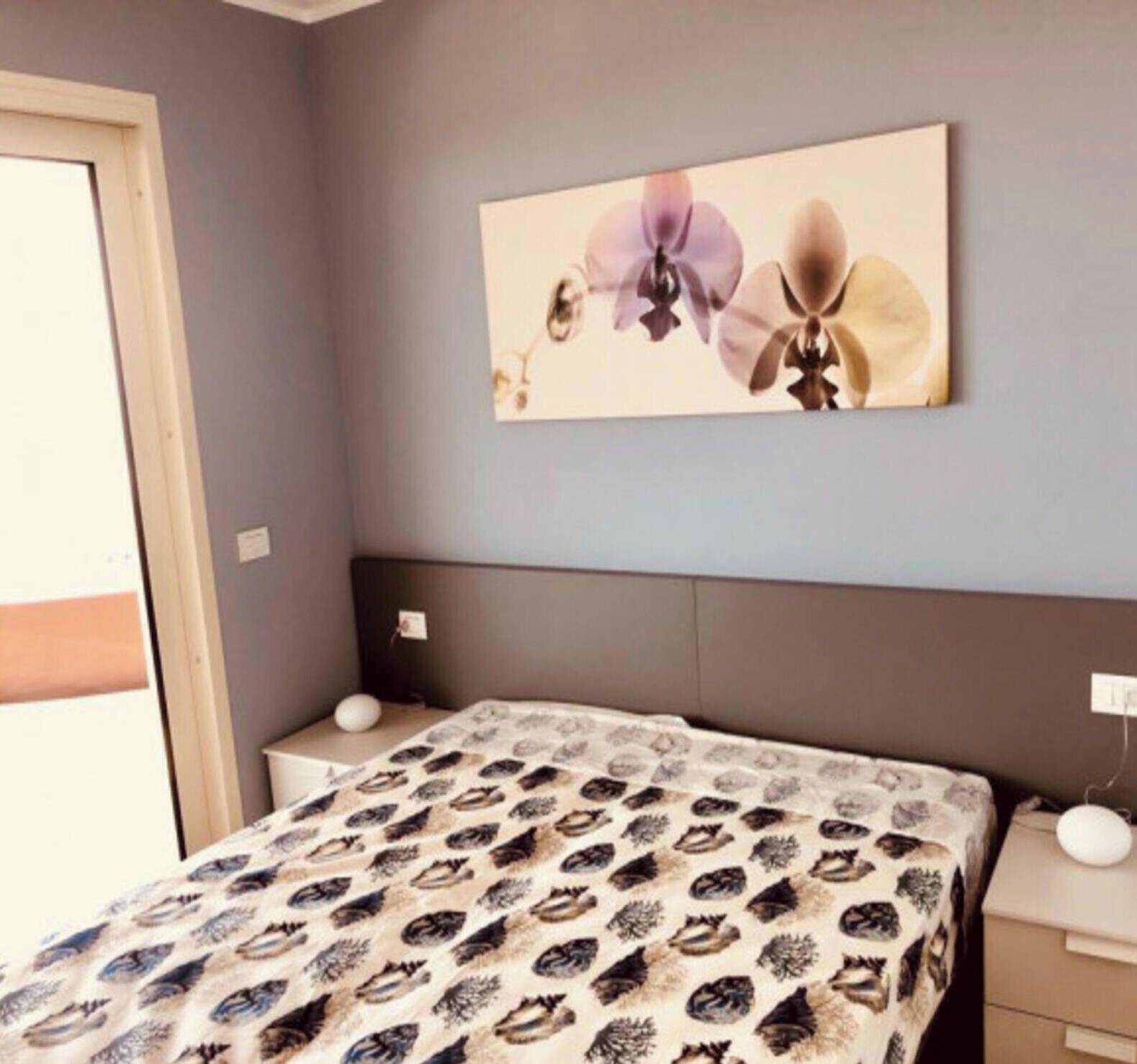 A bedroom with a bed, a nightstand, a wall-mounted lamp, and a floral artwork.