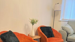 Due poltrone arancioni con cuscini neri, un tavolino rotondo con un vaso di fiori e un divano grigio in una stanza con pavimento tappezzato e una finestra con persiane.