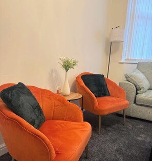 Deux fauteuils orange avec des coussins noirs, une petite table ronde ornée d’un vase de fleurs et un canapé gris dans une pièce dont le sol est recouvert d’une moquette et qui possède une fenêtre avec des stores.