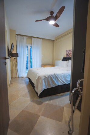 Un dormitorio con cama, ventilador de techo, ventana con cortinas, radiador y mesita de noche.