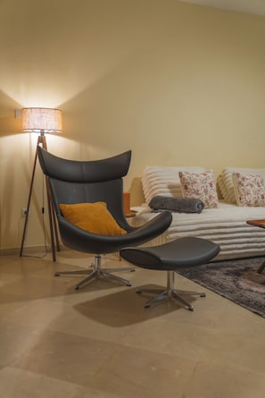Ein modernes Zimmer mit einem geschwungenen schwarzen Sessel, einem kleinen Tisch, einem Bett mit weißer Bettwäsche und floralen Kissen sowie einer Stehlampe.