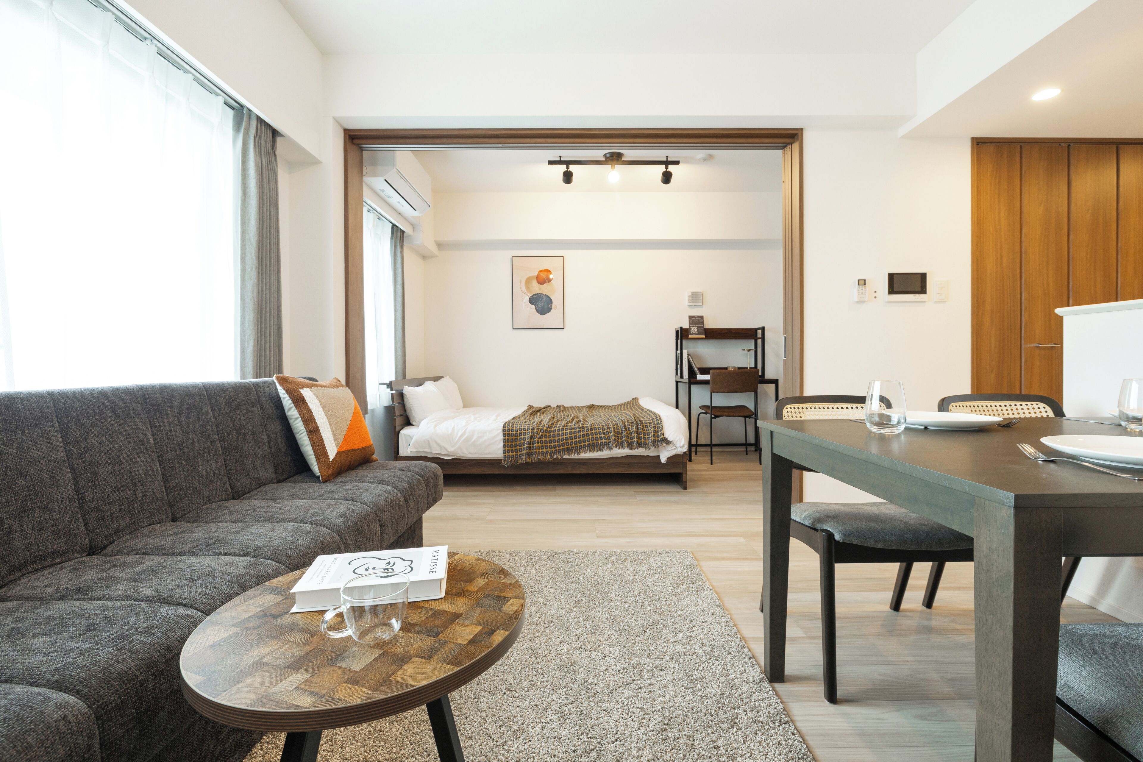 A modern living room with a sofa, a dining table, and a bed in the background.