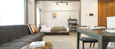 A modern living room with a sofa, a dining table, and a bed in the background.