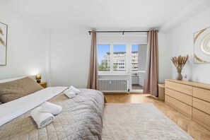 Une chambre à coucher avec un lit, une commode, un radiateur et une vue sur les immeubles par la fenêtre.