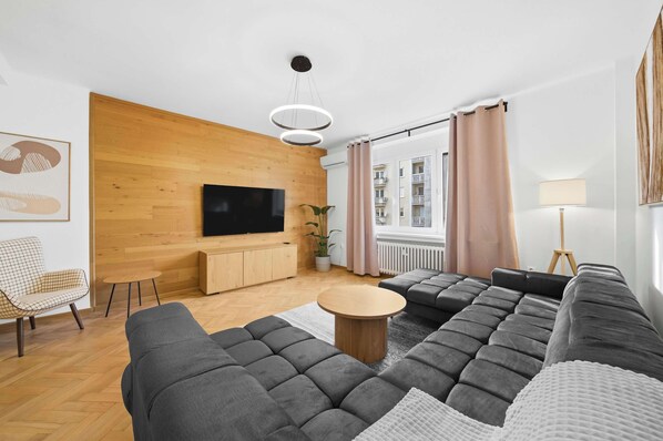 Un salon moderne avec un grand canapé d’angle, une table basse ronde, un téléviseur à écran plat fixé à un mur en bois, une petite table d’appoint, une plante en pot et une vue sur les bâtiments à travers de grandes fenêtres.