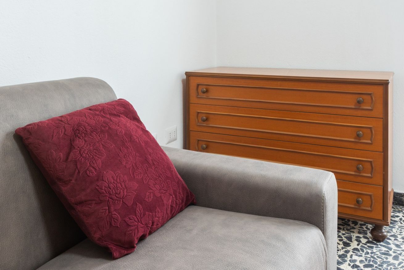Een grijze bank met een rode kussen met bloemen en een houten dressoir met vier lades.