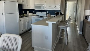 A kitchen with white cabinets, a microwave, a stove, and a bar area with stools. - Beautiful 3BR Condo in OBX, NC, Beach, Pools, Hot Tub, Family Resort Aug. 9-15 (duck)
