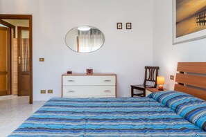 Un dormitorio con cama, cómoda, silla, mesita de noche con lámpara, espejo y un cuadro en la pared.