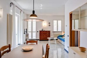 Ein Essbereich mit Tisch, Stühlen, einer Hängeleuchte, einem Holzschrank und einer Couch im Hintergrund.