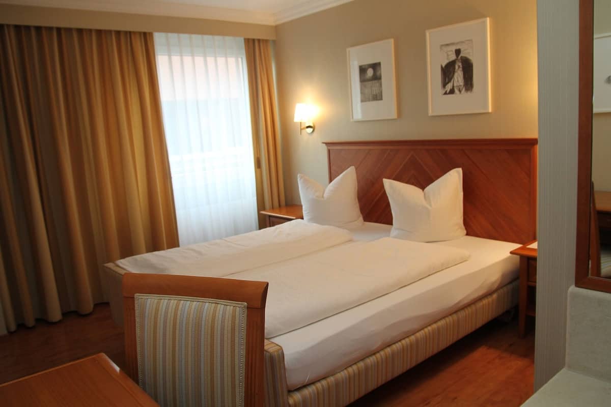 Ein Hotelzimmer mit einem großen Bett, einem Holzkopfstück, einem Nachttisch, einem Sessel und einem Fenster mit Vorhängen.