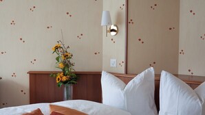 A hotel room with a bed, pillows, a vase with flowers, a wall-mounted lamp, and a wall with a floral pattern.