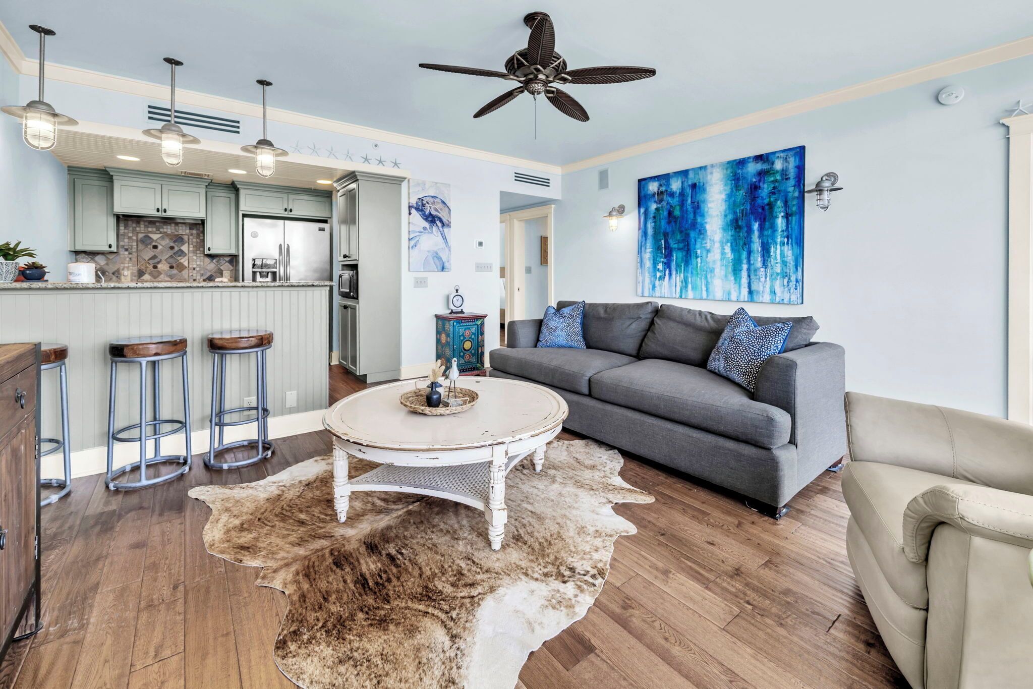 A modern kitchen with bar stools, a dining area with a round table, a gray sofa, and a ceiling fan.