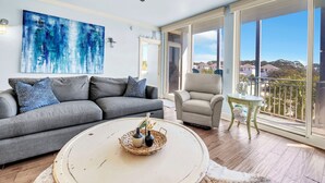 Una sala de estar con un sofá gris, una mesa redonda de centro, un sillón color crema y vistas al exterior a través de ventanas amplias.