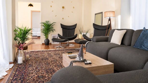 Ein modernes Wohnzimmer mit grauem Sofa, schwarzen Stühlen, einem Holztisch und einem gemusterten Teppich.