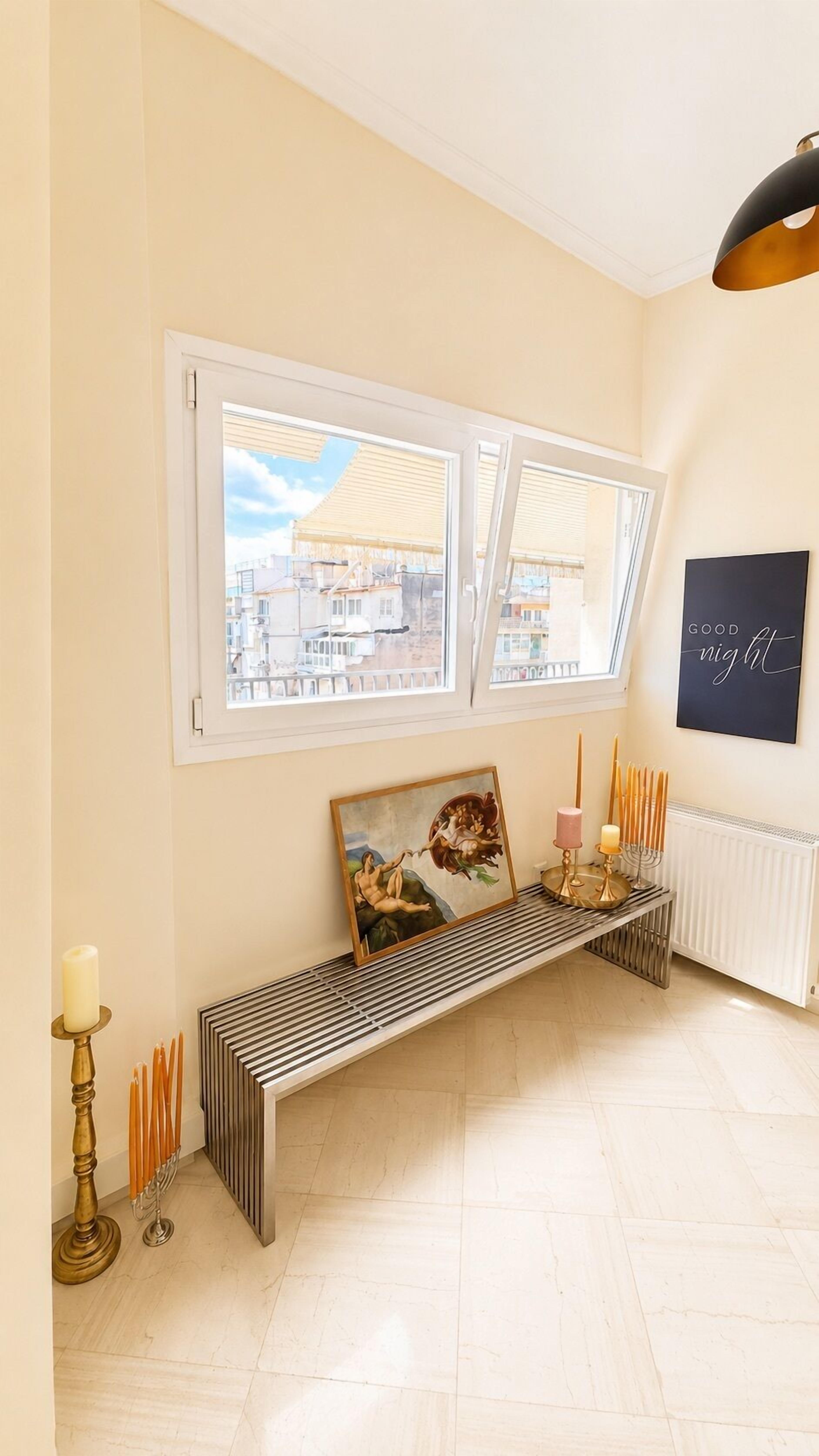 Et værelse med bænk, radiator, et billed, der sidder på væggen, og et vindue med udsigt til bygninger.