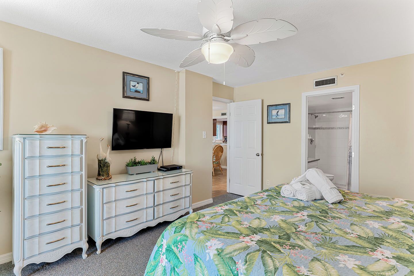 Un dormitorio con cama, televisor, cómoda, ventilador de techo y puerta que da a un baño.