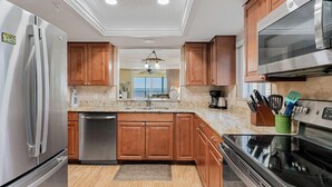 A kitchen with wooden cabinets, stainless steel appliances, a dishwasher, and a countertop with kitchen utensils. - Sand Castle III 205- GULFFRONT CORNER UNIT! (Indian Shores)