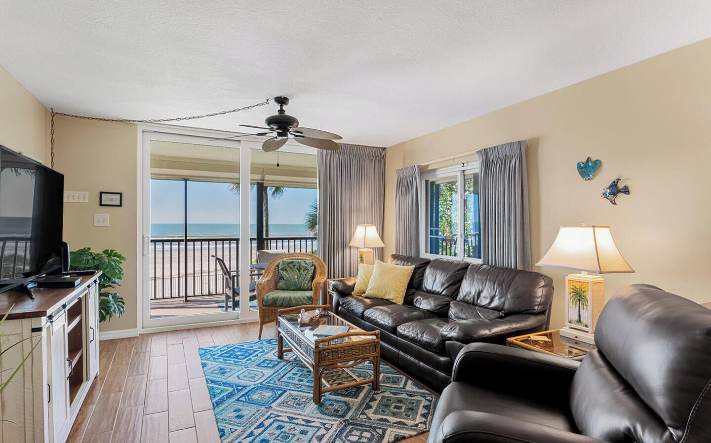 A living room with a leather sofa, a television, a ceiling fan, and a view of the beach.