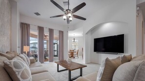 A modern living room with a ceiling fan, a flat-screen TV, and a dining area in the background. - CP948 -6BR  Modern Home | Private Pool | Spacious (Davenport)