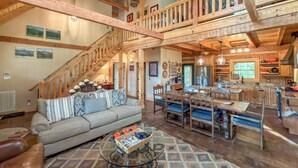 A spacious living area with a high ceiling, wooden beams, a staircase, a sofa, a coffee table, and a dining area with a bench.