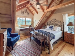 A bedroom with a sloped ceiling, a bed with a metal frame, a desk with a chair, and a window with a view of trees.