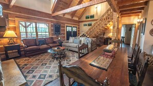 A spacious living area with a high ceiling, wooden beams, a large dining table, and a staircase leading to an upper level.