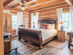 A log cabin-style bedroom with a wooden bed, a ceiling fan, a nightstand, a dresser, and a window with curtains.