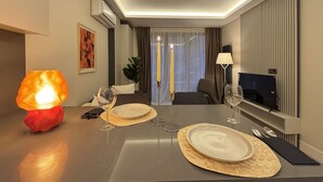 Hotellihuone, jossa on kahdelle katettu ruokapöytä, valaistu lamppu, televisio ja ikkuna, jossa on verhot.