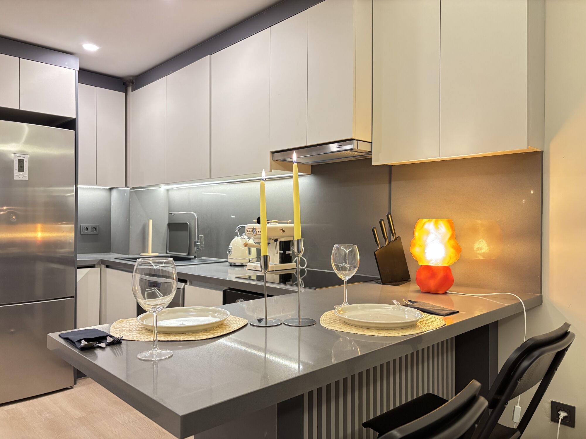 A modern kitchen with a central island, stainless steel appliances, and a dining area with a candle and glassware.