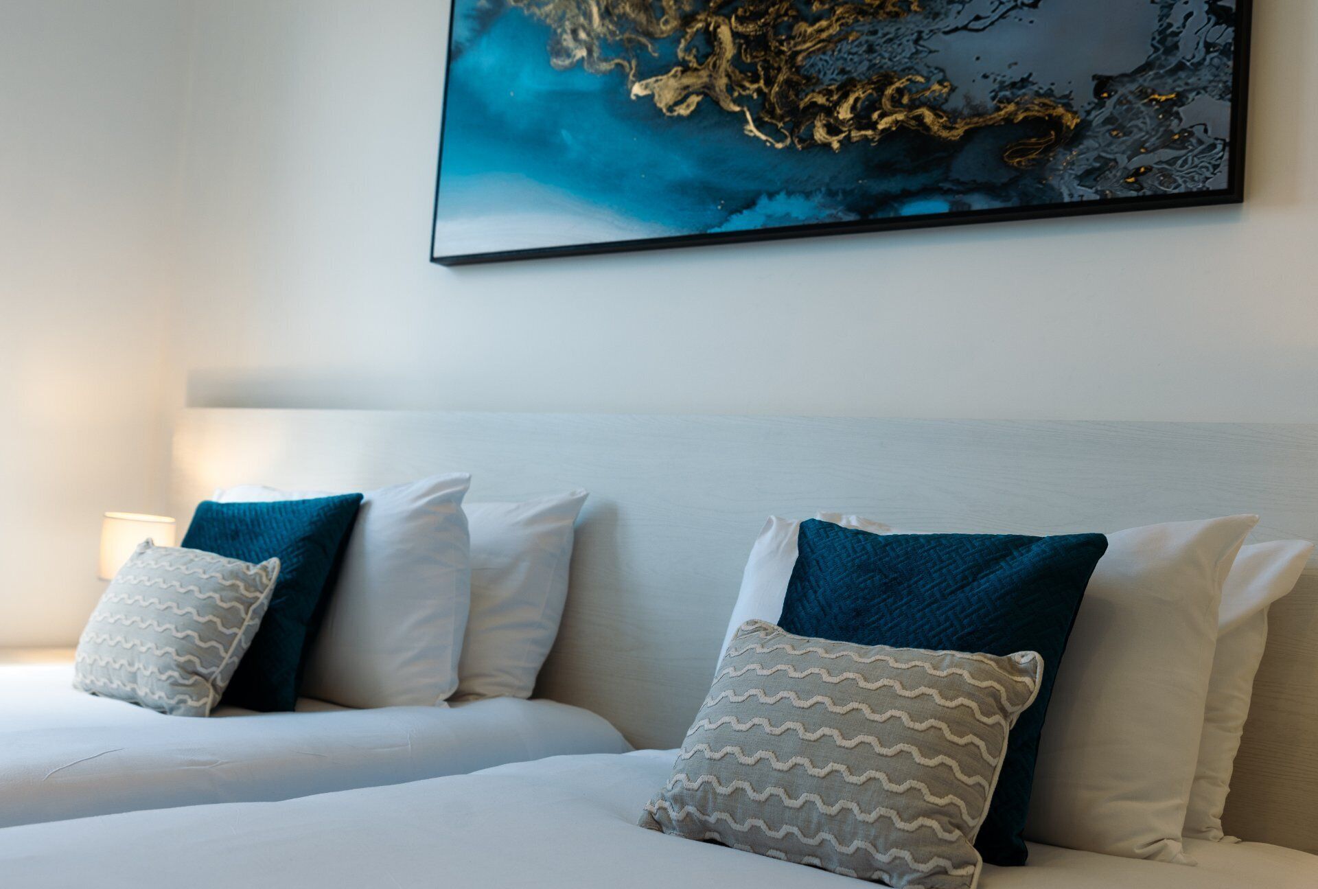 A hotel room with a bed, pillows, a nightstand, and a wall-mounted painting.