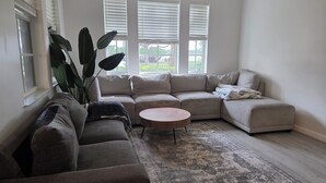 Una sala de estar con un sofá modular, una mesa de centro redonda y ventanas amplias con persianas.