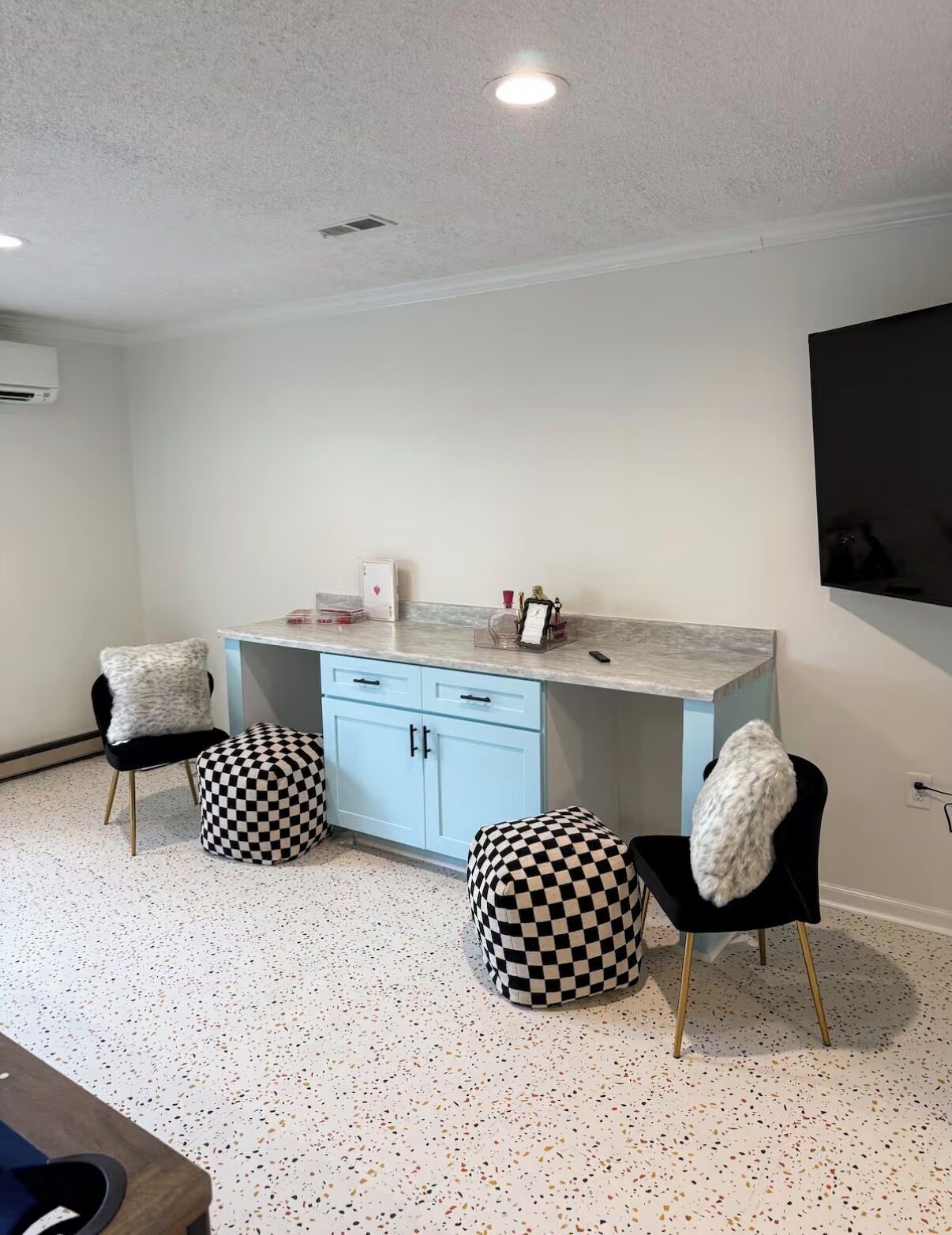 A modern kitchen with a marble countertop, two black chairs with checkered ottomans, a white cabinet, and a mounted flat-screen TV.