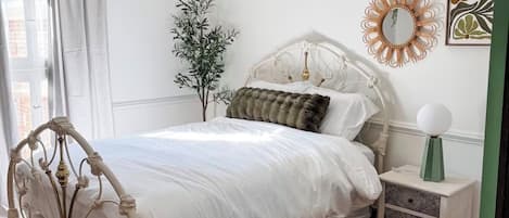 A bedroom with a white bed, a green nightstand, a decorative mirror, and a plant.