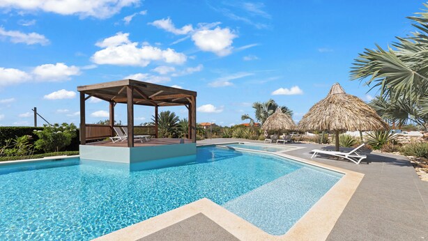 Ein Pool mit einer Holz-Laube, Liegestühlen und reetgedeckten Sonnenschirmen.