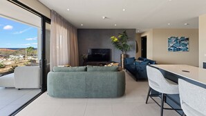 Ein modernes Wohnzimmer mit großem Fenster, Flachbildfernseher, einer Couch, einem Esstisch mit Stühlen und einer Pflanze.