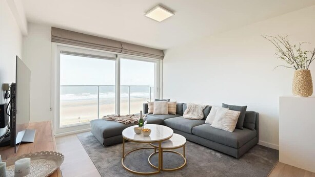 Una sala de estar moderna con un sofá gris, una mesa de centro redonda, un amplio ventanal con vista a la playa y un televisor de pantalla plana.