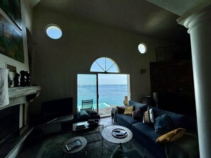 A living room with a large window overlooking the sea, a sofa, a coffee table, and a fireplace. - Villa debut Cap Corse 5mn D'ERBALONGA (BRANDO)