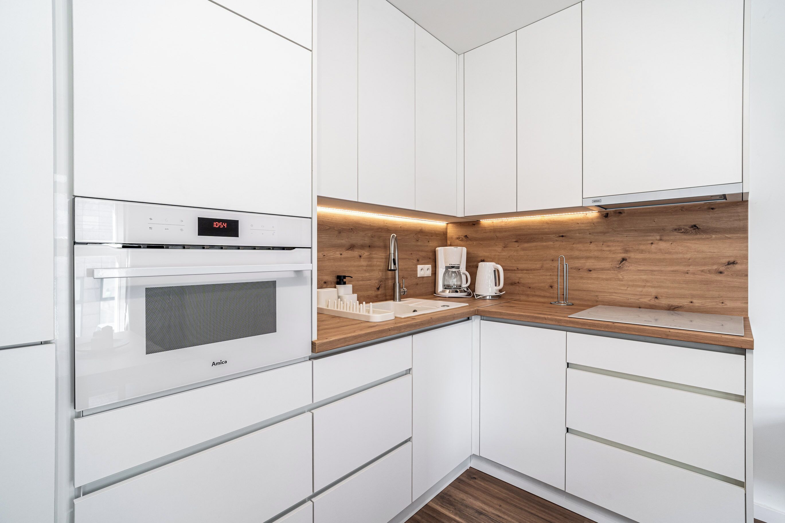 A modern kitchen with white cabinetry, a built-in oven, a coffee maker, and a wooden backsplash.
